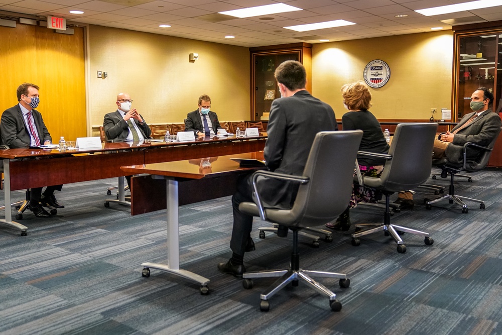 A formal meeting is taking place in what appears to be a USAID conference room, with six individuals seated around tables arranged in a U-shape or parallel configuration. Most participants are wearing face masks, suggesting this occurred during the COVID-19 pandemic period. The USAID seal is visible on the wall, confirming the institutional setting.