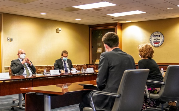 A formal meeting is taking place in what appears to be a USAID conference room, with six individuals seated around tables arranged in a U-shape or parallel configuration. Most participants are wearing face masks, suggesting this occurred during the COVID-19 pandemic period. The USAID seal is visible on the wall, confirming the institutional setting.
