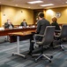 A formal meeting is taking place in what appears to be a USAID conference room, with six individuals seated around tables arranged in a U-shape or parallel configuration. Most participants are wearing face masks, suggesting this occurred during the COVID-19 pandemic period. The USAID seal is visible on the wall, confirming the institutional setting.