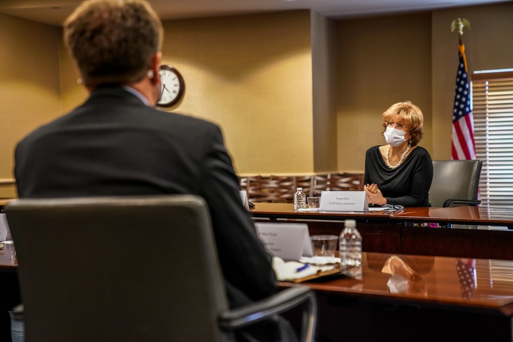 A formal meeting is taking place in a conference room between at least two individuals seated at a large wooden table. The woman facing the camera is wearing a white face mask and a black outfit, with a name placard in front of her. An American flag is visible in the background, suggesting an official or governmental setting.