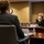 A formal meeting is taking place in a conference room between at least two individuals seated at a large wooden table. The woman facing the camera is wearing a white face mask and a black outfit, with a name placard in front of her. An American flag is visible in the background, suggesting an official or governmental setting.
