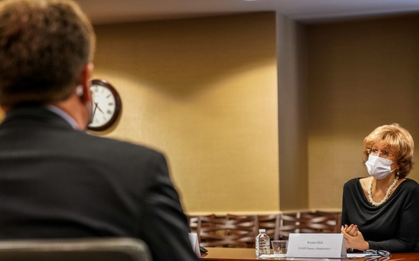 A formal meeting is taking place in a conference room between at least two individuals seated at a large wooden table. The woman facing the camera is wearing a white face mask and a black outfit, with a name placard in front of her. An American flag is visible in the background, suggesting an official or governmental setting.