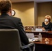 A formal meeting is taking place in a conference room between at least two individuals seated at a large wooden table. The woman facing the camera is wearing a white face mask and a black outfit, with a name placard in front of her. An American flag is visible in the background, suggesting an official or governmental setting.