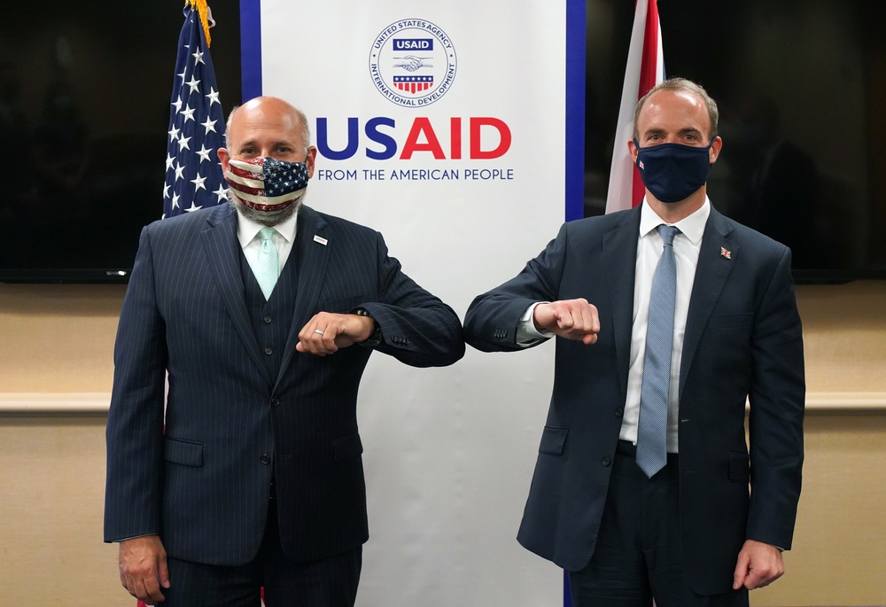 Two men in dark suits are performing an elbow bump greeting in front of a USAID (United States Agency for International Development) banner. Both individuals are wearing face masks, suggesting this photo was taken during the COVID-19 pandemic period. An American flag and what appears to be a Union Jack flag are visible in the background.