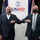 Two men in dark suits are performing an elbow bump greeting in front of a USAID (United States Agency for International Development) banner. Both individuals are wearing face masks, suggesting this photo was taken during the COVID-19 pandemic period. An American flag and what appears to be a Union Jack flag are visible in the background.