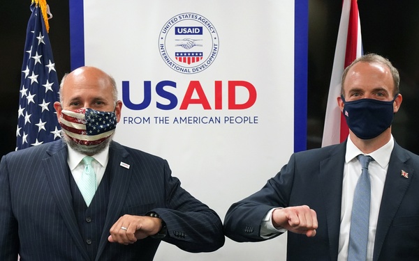 Two men in dark suits are performing an elbow bump greeting in front of a USAID (United States Agency for International Development) banner. Both individuals are wearing face masks, suggesting this photo was taken during the COVID-19 pandemic period. An American flag and what appears to be a Union Jack flag are visible in the background.