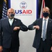Two men in dark suits are performing an elbow bump greeting in front of a USAID (United States Agency for International Development) banner. Both individuals are wearing face masks, suggesting this photo was taken during the COVID-19 pandemic period. An American flag and what appears to be a Union Jack flag are visible in the background.