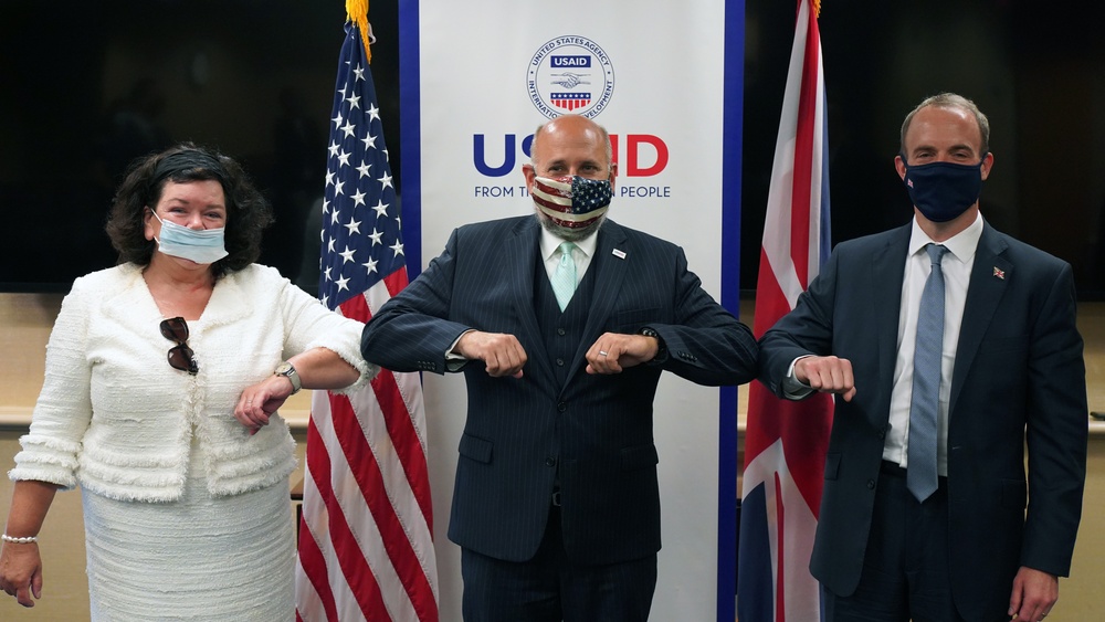 Three individuals wearing face masks are performing an elbow bump greeting in front of a USAID banner and flags of the United States and United Kingdom. The setting appears to be an official diplomatic or governmental event, indicated by the institutional branding and national flags visible in the background. All three participants are dressed in formal attire.