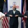Three individuals wearing face masks are performing an elbow bump greeting in front of a USAID banner and flags of the United States and United Kingdom. The setting appears to be an official diplomatic or governmental event, indicated by the institutional branding and national flags visible in the background. All three participants are dressed in formal attire.