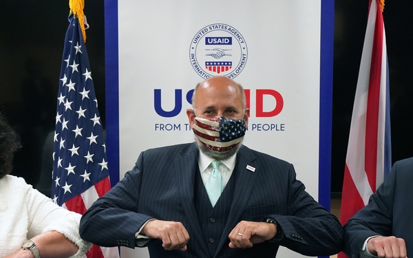 Three individuals wearing face masks are performing an elbow bump greeting in front of a USAID banner and flags of the United States and United Kingdom. The setting appears to be an official diplomatic or governmental event, indicated by the institutional branding and national flags visible in the background. All three participants are dressed in formal attire.