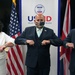 Three individuals wearing face masks are performing an elbow bump greeting in front of a USAID banner and flags of the United States and United Kingdom. The setting appears to be an official diplomatic or governmental event, indicated by the institutional branding and national flags visible in the background. All three participants are dressed in formal attire.