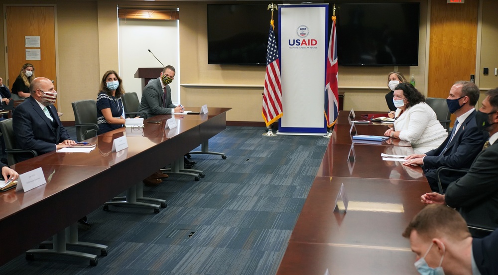 A formal bilateral meeting is taking place at USAID headquarters, indicated by the prominent USAID banner and the presence of both an American flag and a British Union Jack flag. Participants are seated on opposite sides of a large conference table, with most wearing face masks suggesting this occurred during the COVID-19 pandemic period. The setting appears to be an official diplomatic or aid coordination meeting between US and UK representatives.