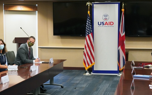 A formal bilateral meeting is taking place at USAID headquarters, indicated by the prominent USAID banner and the presence of both an American flag and a British Union Jack flag. Participants are seated on opposite sides of a large conference table, with most wearing face masks suggesting this occurred during the COVID-19 pandemic period. The setting appears to be an official diplomatic or aid coordination meeting between US and UK representatives.