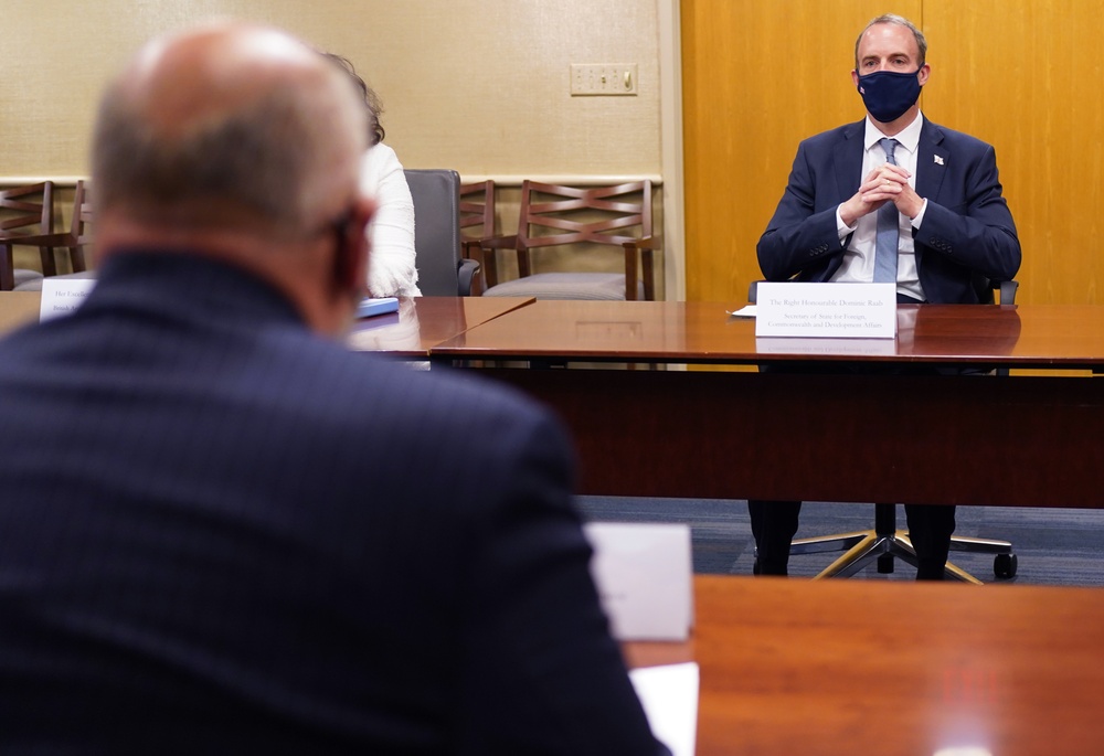 A formal meeting is taking place between at least two individuals seated at a large conference table. The man facing the camera is wearing a dark suit and a dark face mask, with a nameplate in front of him. The setting appears to be an official diplomatic or governmental meeting room.