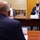 A formal meeting is taking place between at least two individuals seated at a large conference table. The man facing the camera is wearing a dark suit and a dark face mask, with a nameplate in front of him. The setting appears to be an official diplomatic or governmental meeting room.