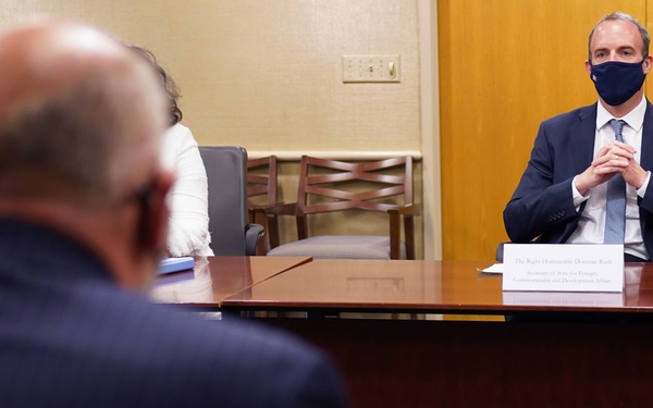 A formal meeting is taking place between at least two individuals seated at a large conference table. The man facing the camera is wearing a dark suit and a dark face mask, with a nameplate in front of him. The setting appears to be an official diplomatic or governmental meeting room.