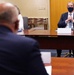 A formal meeting is taking place between at least two individuals seated at a large conference table. The man facing the camera is wearing a dark suit and a dark face mask, with a nameplate in front of him. The setting appears to be an official diplomatic or governmental meeting room.