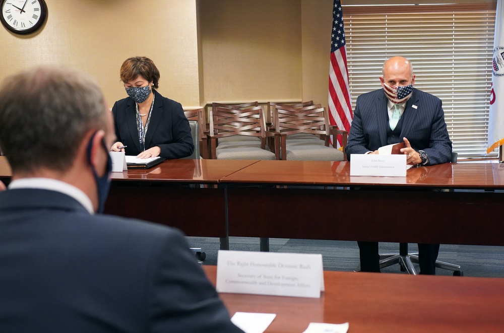 A formal meeting is taking place in a conference room with participants wearing face masks, suggesting a COVID-19 era setting. Three individuals are seated around a large wooden conference table with name placards visible. An American flag and what appears to be an organizational seal or flag are visible in the background.