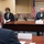 A formal meeting is taking place in a conference room with participants wearing face masks, suggesting a COVID-19 era setting. Three individuals are seated around a large wooden conference table with name placards visible. An American flag and what appears to be an organizational seal or flag are visible in the background.