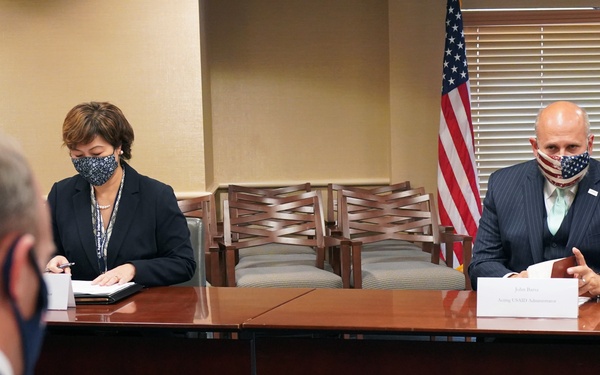 A formal meeting is taking place in a conference room with participants wearing face masks, suggesting a COVID-19 era setting. Three individuals are seated around a large wooden conference table with name placards visible. An American flag and what appears to be an organizational seal or flag are visible in the background.