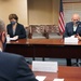 A formal meeting is taking place in a conference room with participants wearing face masks, suggesting a COVID-19 era setting. Three individuals are seated around a large wooden conference table with name placards visible. An American flag and what appears to be an organizational seal or flag are visible in the background.