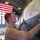 A military or civilian technician is applying what appears to be a decal or stencil featuring an American flag design onto a large gray aircraft surface inside a hangar. The work is being performed in a spacious military hangar, evidenced by the large American flag hanging in the background and partial text reading 'U.S.A.F.' on the aircraft. The number '42' is also visible on the aircraft fuselage.