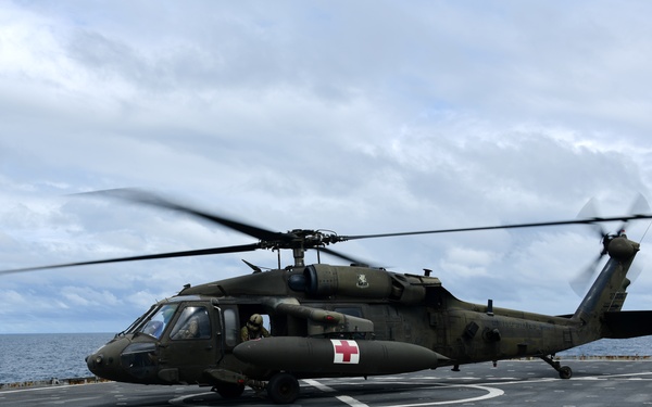 A military medical evacuation (MEDEVAC) helicopter, identifiable by the red cross markings on its fuselage, is landed on the flight deck of a naval vessel at sea. The helicopter appears to be an Army UH-60 Black Hawk variant configured for medical operations, with its rotors spinning. The scene takes place over open ocean under a partly cloudy sky.