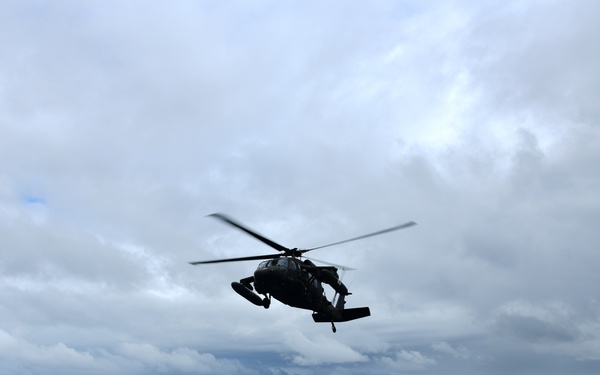 A military helicopter, consistent with a UH-60 Black Hawk silhouette, is hovering or landing/departing over the flight deck of a naval vessel at sea. The ship's deck features marked landing zones and safety equipment visible along the edges. The scene takes place under an overcast, cloudy sky over open ocean.