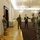 A formal meeting or reception is taking place in an ornate government or official building. A civilian in a suit is engaging with several military personnel wearing camouflage uniforms and face masks. The setting features decorative chandeliers, parquet flooring, and what appears to be an official coat of arms on the wall.