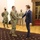 A formal meeting is taking place in what appears to be an official government or military building. A US Army soldier in OCP (Operational Camouflage Pattern) uniform is shaking hands with a civilian man in a navy blue suit. All individuals present are wearing face masks, suggesting this occurred during the COVID-19 pandemic period.