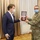 A formal presentation ceremony showing two individuals exchanging what appears to be a commemorative medal or plaque in a velvet-lined display box. One person is wearing a blue civilian suit while the other is dressed in U.S. Army camouflage (OCP) uniform with a 'MED' patch. Both individuals are wearing face masks, suggesting the event took place during the COVID-19 pandemic period.