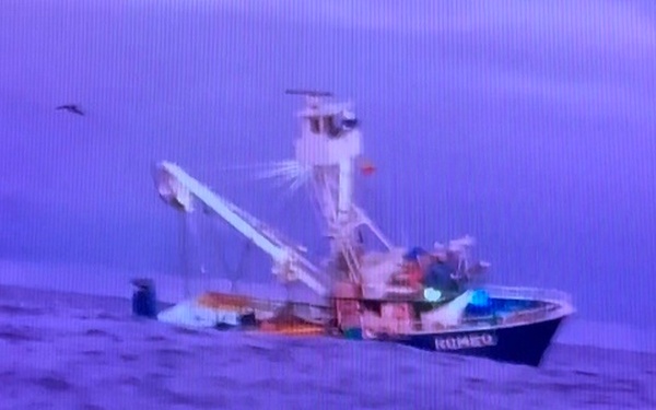 A commercial fishing vessel, appearing to be named 'Romeo', is photographed at sea during low-light conditions, likely dusk or dawn. The boat is equipped with outriggers, booms, and rigging typical of a shrimp trawler or similar commercial fishing craft. The image appears to be a photograph of a screen, resulting in a purple-blue color cast and visible scan lines.