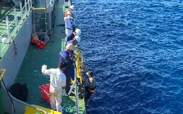 The image shows a maritime rescue or transfer operation taking place alongside a large vessel with a green-painted deck. Several crew members are positioned along the ship's railing, appearing to assist people in an orange inflatable rescue boat below. The scene takes place in open blue ocean waters under daylight conditions.