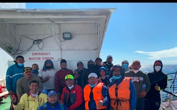 A group of approximately 15 individuals posing for a group photo on the deck of a vessel near an 'Emergency Muster Station' sign. Several people are wearing face masks and one person in the center is wearing an orange life vest. The ocean is visible in the background under a clear blue sky.