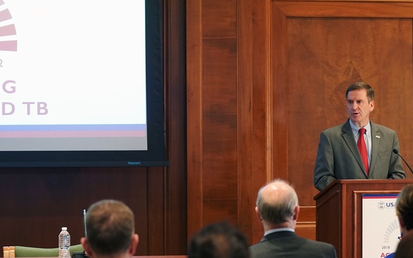 A man in a gray suit and red tie is delivering a speech at a wooden podium bearing a USAID logo during what appears to be a tuberculosis-related conference or event. A large projection screen behind him displays a presentation slide reading '2022 ACCELERATING [EFFORTS] TO END TB.' An audience of approximately 8-10 attendees is seated facing the speaker.