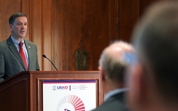 A man in a gray suit and red tie is speaking at a wooden podium during a formal event. The podium displays a USAID and National Academies of Sciences, Engineering, and Medicine co-branded sign referencing a 2018–2022 initiative titled 'Accelerating Progress Toward...' An audience is visible in the foreground, with one attendee recording on a smartphone.