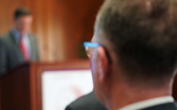 A man in a dark suit is using a smartphone to livestream or record a speaker at a podium in what appears to be a formal indoor venue. The phone screen shows a 'LIVE' indicator, suggesting the event is being broadcast in real time. The background features warm wood paneling and a blurred figure of the speaker wearing a gray suit and red tie.