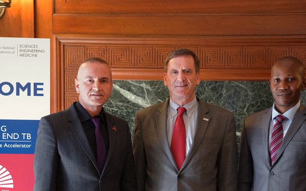 Three men in business attire pose for a formal photograph at a USAID event titled 'Accelerating Progress to End TB - First Anniversary of the Accelerator,' held on November 26, 2019. The event is co-sponsored by USAID and the National Academies of Sciences, Engineering, and Medicine. The setting appears to be an official government or institutional building with wood-paneled walls and marble accents.