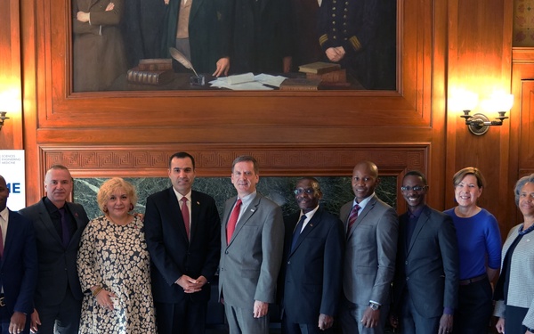 A formal group photo of approximately 11 individuals standing in a wood-paneled conference or boardroom setting. A USAID banner is visible in the background along with a large oil painting. The gathering appears to be an official meeting or delegation event.