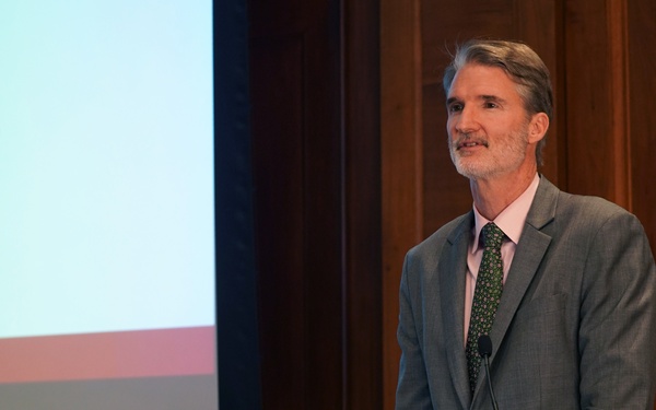 A middle-aged man with gray hair and a short beard is speaking at a wooden podium during a formal conference or event. The podium displays a sign indicating a USAID and National Academies of Sciences, Engineering, and Medicine joint event titled 'Accelerating Progress to End TB' spanning 2018–2022. A presentation screen is partially visible in the background.
