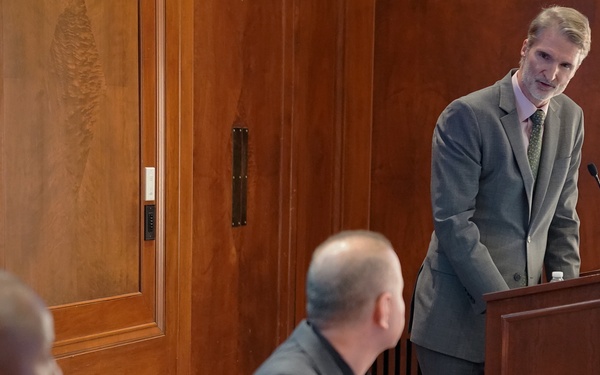 A formal meeting or hearing is taking place in a wood-paneled room, likely a government or institutional setting. A tall man in a gray suit stands at a wooden podium speaking, while two other men in suits are seated at a conference table in the foreground. The setting suggests an official briefing, panel discussion, or legislative hearing.