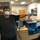 A man wearing an N95 respirator mask and blue latex gloves stands at what appears to be a reception or security desk, pointing toward something off-frame. The setting appears to be a government or military facility, specifically associated with the Electro-Optic Technology Division. A sign on the desk reads 'Guest Masks,' suggesting this was taken during a period requiring protective equipment protocols.