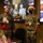 Two military personnel in camouflage uniforms are celebrating Christmas in what appears to be an office or briefing room. One wears a festive ugly Christmas sweater and Santa headband, while the other wears a Santa hat, both wearing black face masks. The setting includes British and American flags, Union Jack decorations, milk cartons, and a monitor displaying a Christmas photo of an elderly woman in a formal setting.
