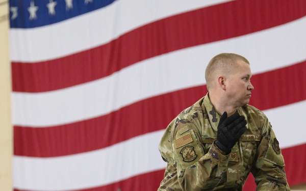A U.S. military service member in OCP (Operational Camouflage Pattern) uniform is crouching on a metal structure inside what appears to be a military hangar or large indoor facility. A large American flag is prominently displayed in the background. The soldier is wearing black tactical gloves and has a unit patch and U.S. flag patch visible on the left sleeve.