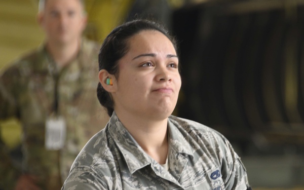 A U.S. Air Force female Airman in Airman Battle Uniform (ABU) is seated in what appears to be an aircraft hangar, looking upward with a contemplative expression. She wears foam earplugs and has a name tape partially visible reading 'TINOCOT' or similar. Additional military personnel in OCP uniforms are visible in the blurred background.