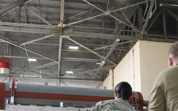 Two military personnel in U.S. Air Force uniforms are working inside a large hangar facility. A large red and gray cylindrical object, consistent with a training or inert munition/weapon component on a loading cart, is prominently visible. The setting appears to be a weapons loading or maintenance training exercise.