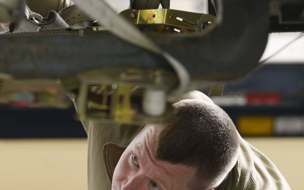 A military technician or mechanic is working underneath what appears to be an aircraft or military vehicle, looking upward to inspect or service equipment. The individual is wearing an olive/tan flight suit and has colorful foam ear protection inserted in their ear. The setting appears to be an aircraft hangar based on the background environment.
