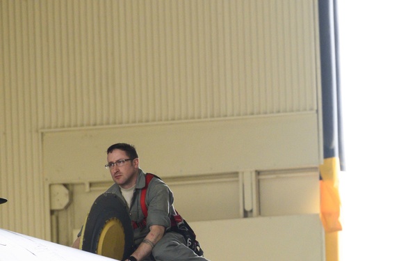 A male technician or mechanic is positioned on top of a large military or commercial aircraft inside a hangar. He appears to be performing an inspection or maintenance task near what looks like a large wheel or engine component. The setting is an aircraft maintenance hangar with yellow structural elements visible in the background.