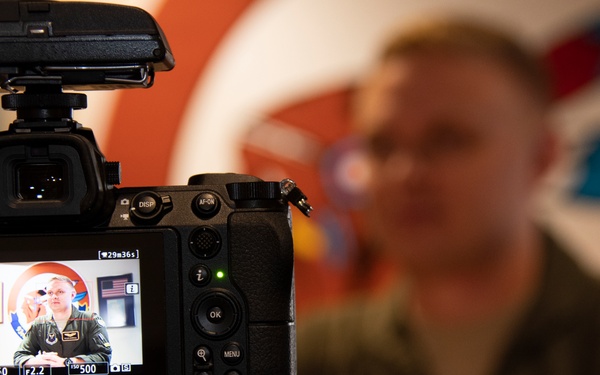 A Nikon mirrorless camera on a tripod is actively recording a military personnel member, as indicated by the red REC indicator on the LCD screen. The subject, wearing an olive/flight suit uniform with patches, is seated in front of a colorful background with what appears to be military insignia or artwork. The camera's LCD display shows the live feed with settings including f/2.2, ISO 500, and approximately 29 minutes 36 seconds of recording time remaining.