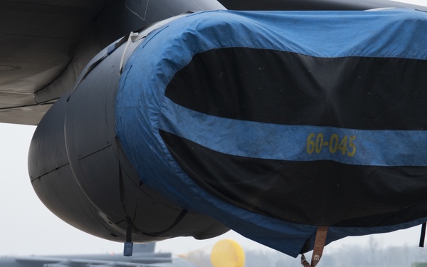 Close-up view of a military aircraft engine nacelle fitted with a protective inlet cover, marked with the identifier '60-045' in yellow stenciling. The cover is blue and black fabric, secured around the engine intake to protect it from foreign object debris and weather while parked. In the background, another large military aircraft (likely a B-52) is visible on the flight line.